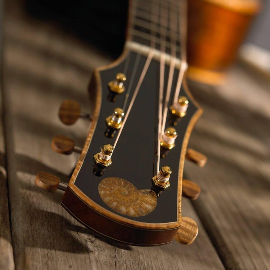 Close-up of a guitar headstock on a wooden surface, featuring a polished black finish, golden tuning pegs, and a fossil shell inlay, with a warm, elegant ambiance.