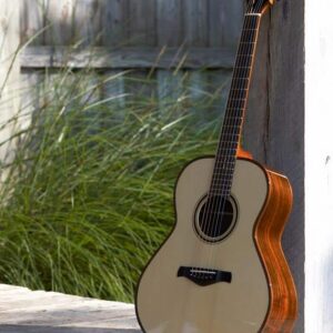 An acoustic guitar leaning against a white wooden post on an outdoor deck, with tall green grass in the background.