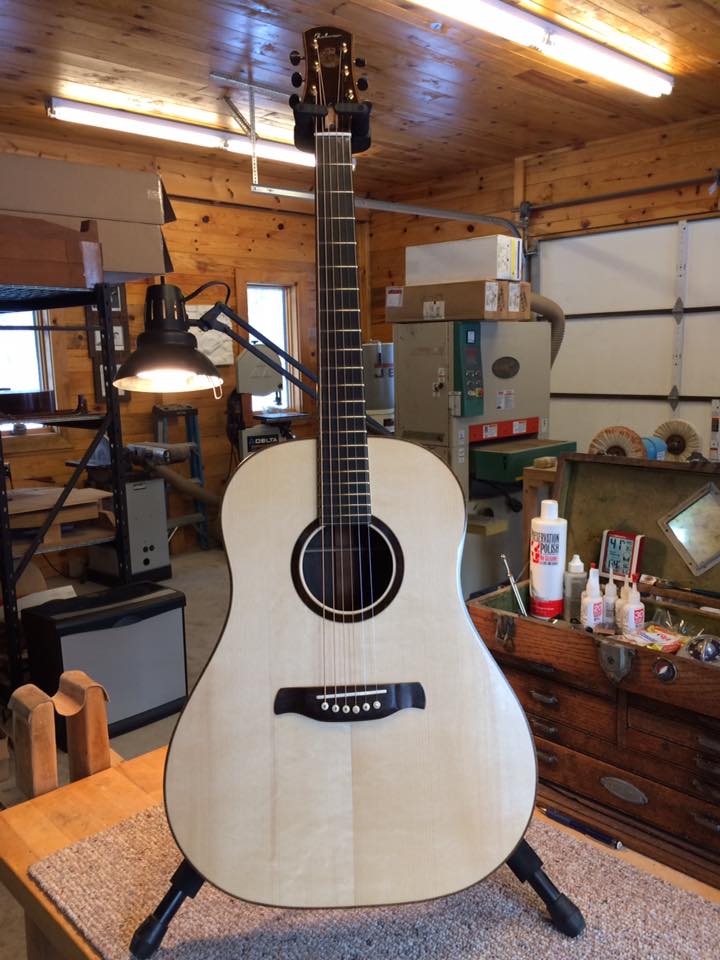 Acoustic guitar with light wood finish on stand in workshop. Surroundings include tools, cabinets, and wood paneling, conveying craftsmanship.