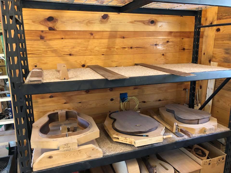 A metal shelving unit in a wood-paneled workshop holding various raw guitar parts and bodies in early stages of construction.