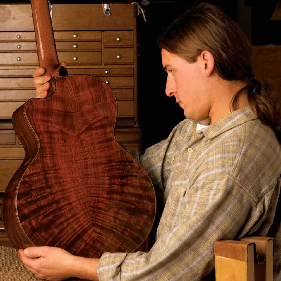A man with long hair inspects a polished, richly textured acoustic guitar body. He wears a plaid shirt and sits in a workshop filled with wooden drawers.
